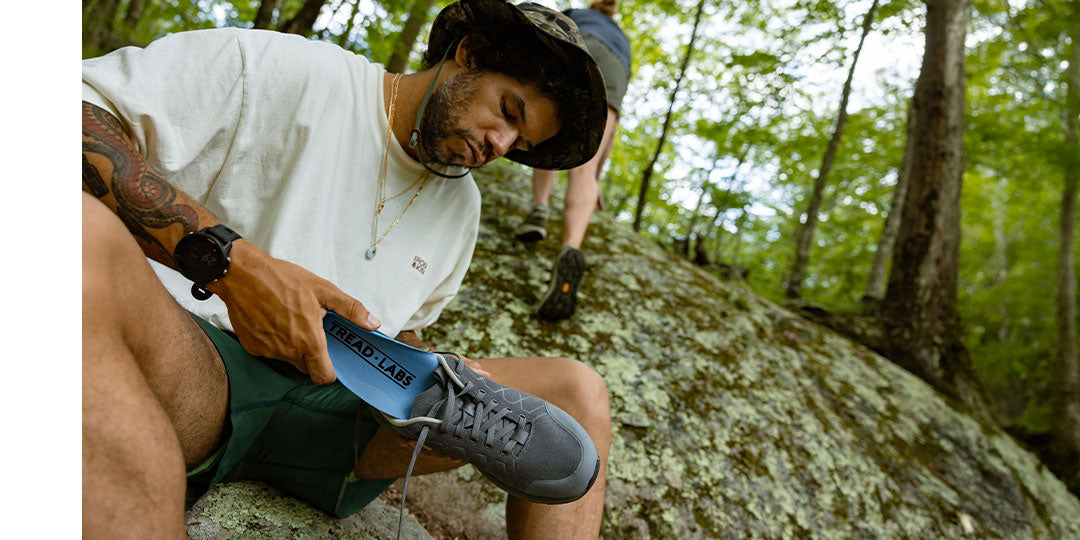 Man adjusting hiking shoes on a rocky trail in a forest