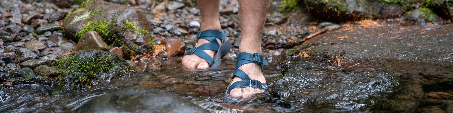 Man wearing blue Redway Sandals stepping into a rocky stream with mossy rocks nearby