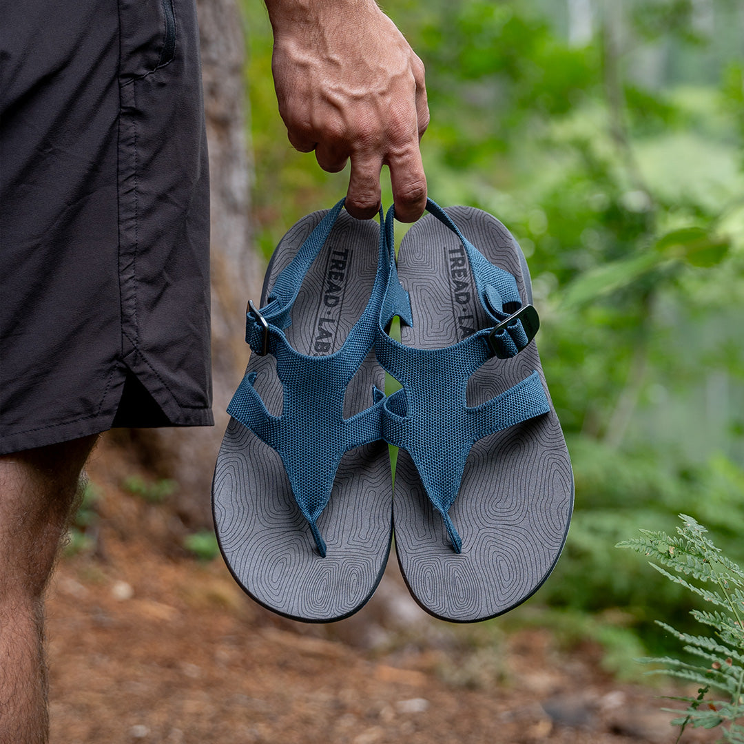 Man holding a pair of blue Albion Sandals in his hands while walking on a hiking trail in the forest