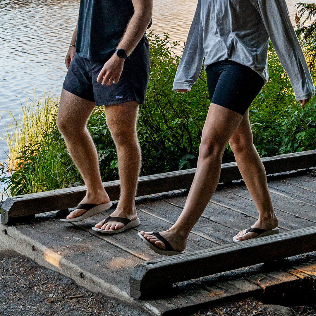 Man and woman each wearing brown leather clip flops from Tread Labs walking on a boardwalk next to a scenic lake
