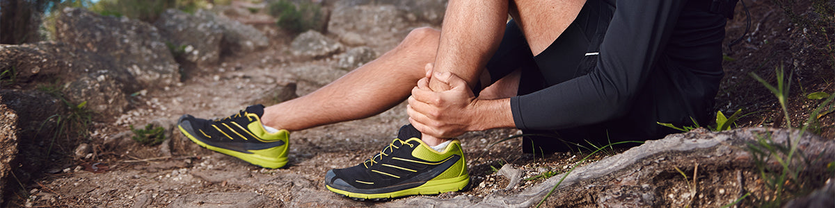 A man suffering from tendonitis clutching his lower leg sitting by some rocks in the forest