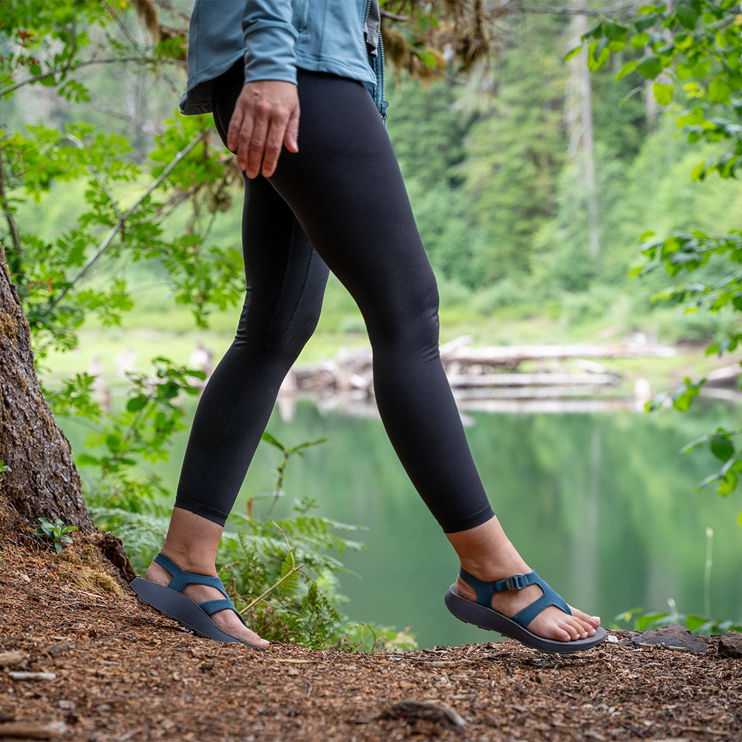 Woman wearing black leggings and blue Albion Sandals with arch support walking through the forest nearby a green lake