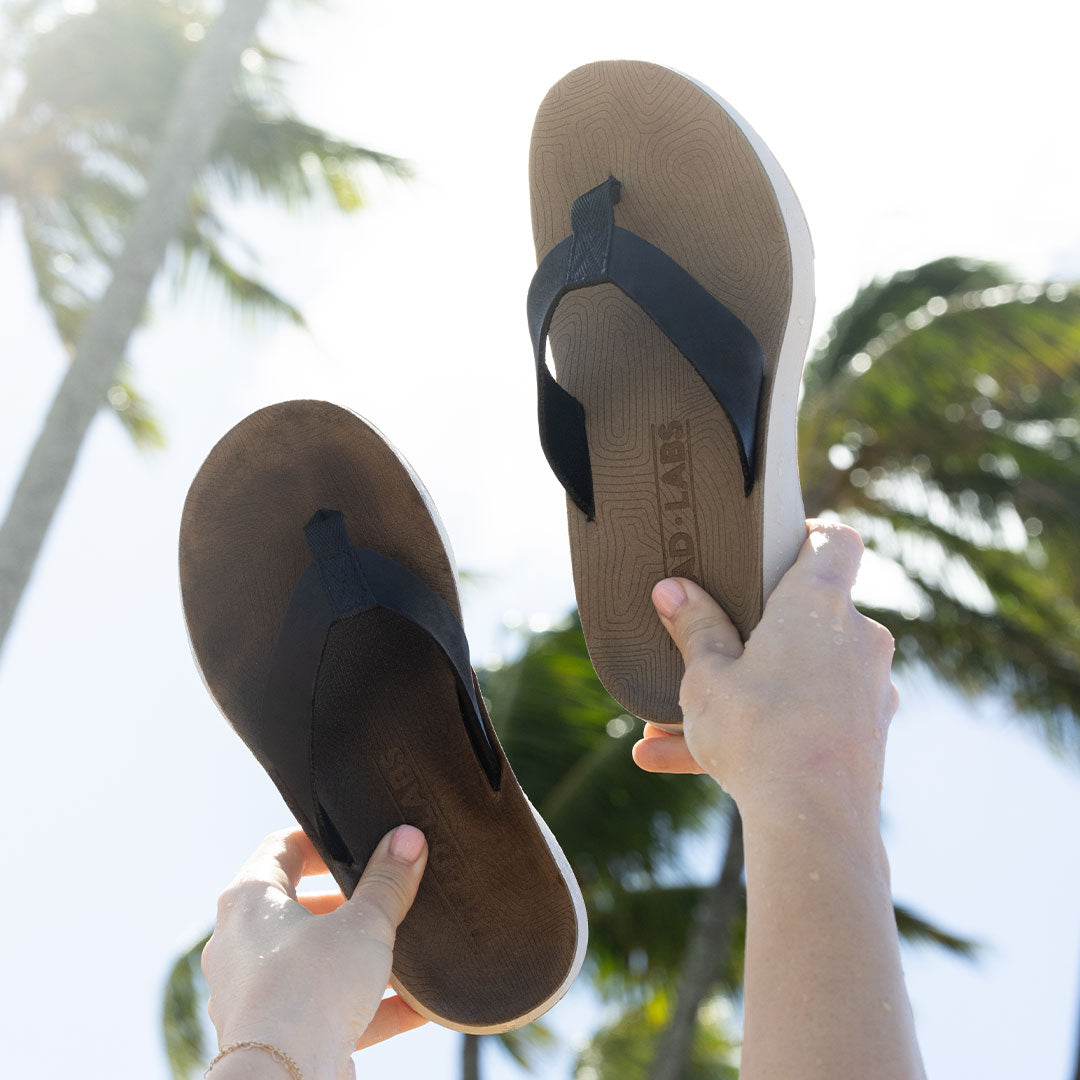 Woman holding brown leather flip flops from Tread Labs in the air in front of palm trees