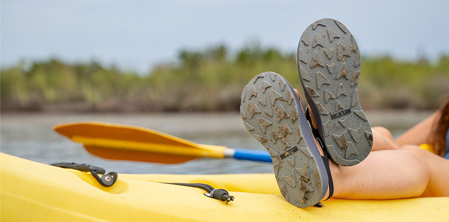 Feet crossed on top of a kayak with the soles of the sandals showing.