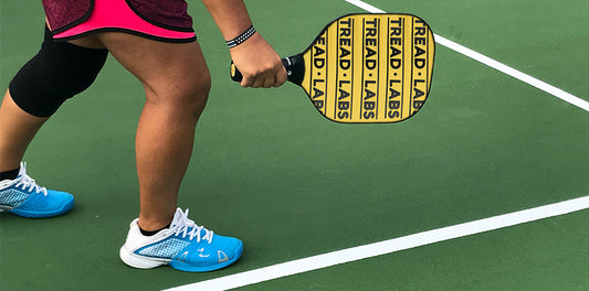 Pickleball player from the waist down wearing turquoise shoes, red shorts, with a yellow paddle on a green court.