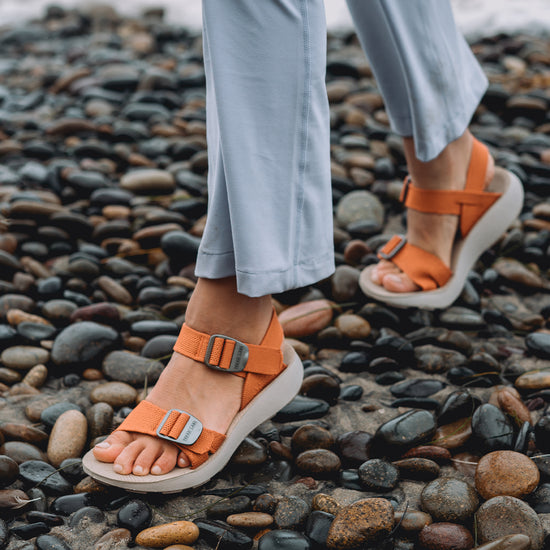 Woman wearing orange Salinas Sandals and blue yoga pants walking on a rocky beach.
