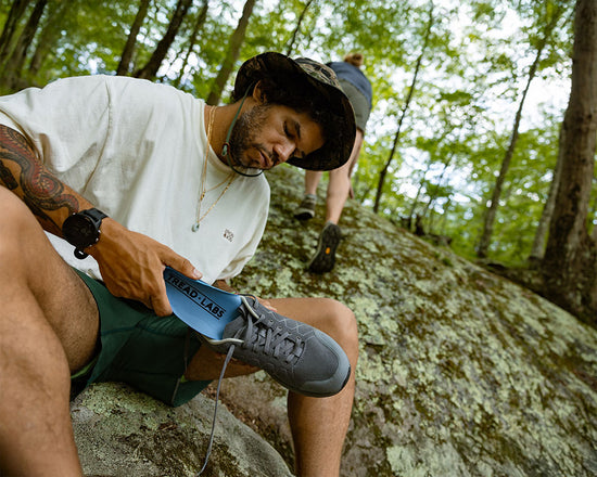 Two people on a rocky path in a forest, one holding a blue trail shoe.