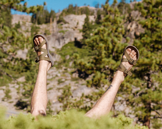 Man wearing Redway Slide sandals putting his feet up into the air with a rocky, forested cliff in the background.