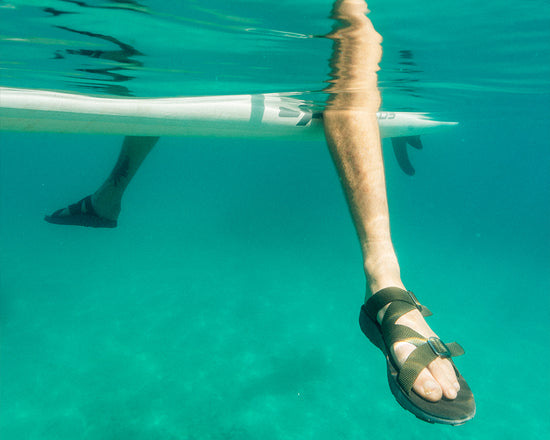 Underwater shot of man wearing Redway Slide Sandals while sitting on a paddleboard in a clear blue lake.