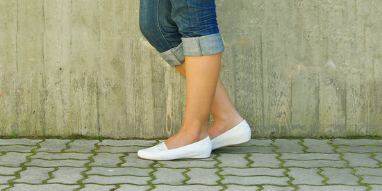 Woman wearing jeans and white dress flats walking on a concrete sidewalk.