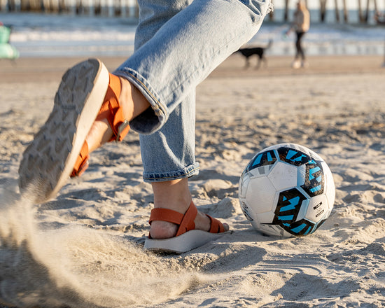 Woman wearing Salinas Sandals in Apricot Orange Color Kicking a Soccer Ball on the Beach