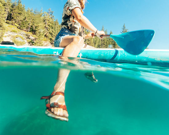 Woman wearing jean shorts and Salinas Slide Sandals sitting on a paddleboard with an underwater view of her sandals.