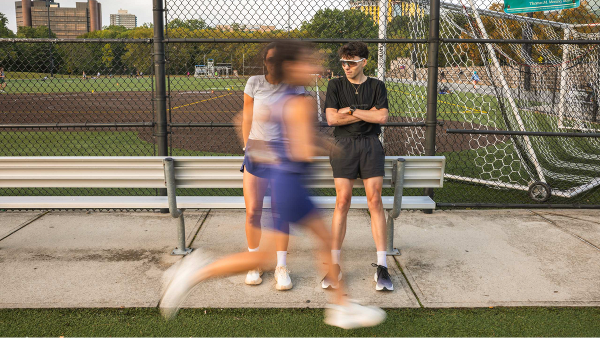 Three people at an outdoor sports field behind a chain-link fence, with one person in the foreground running past in motion blur and two standing behind on a bench.