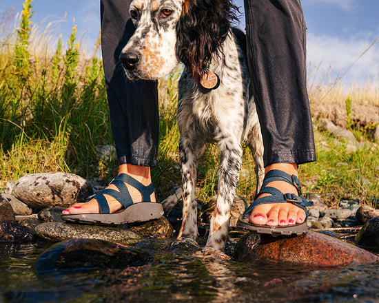 Woman wearing blue Redway sandals standing on a riverbank with her dog.