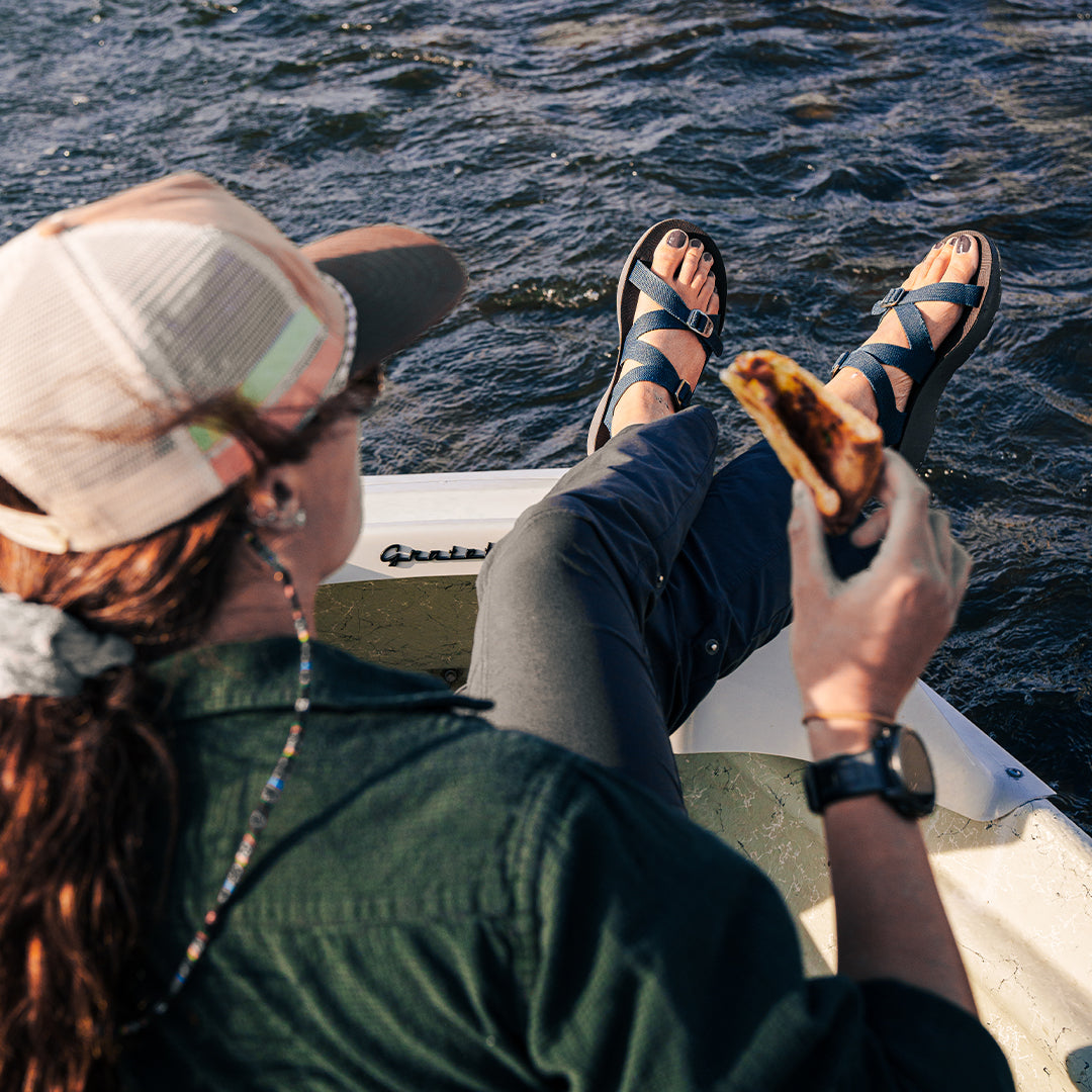 Woman wearing blue Redway Sandals sitting on a fishing boat eating a slice of pizza.